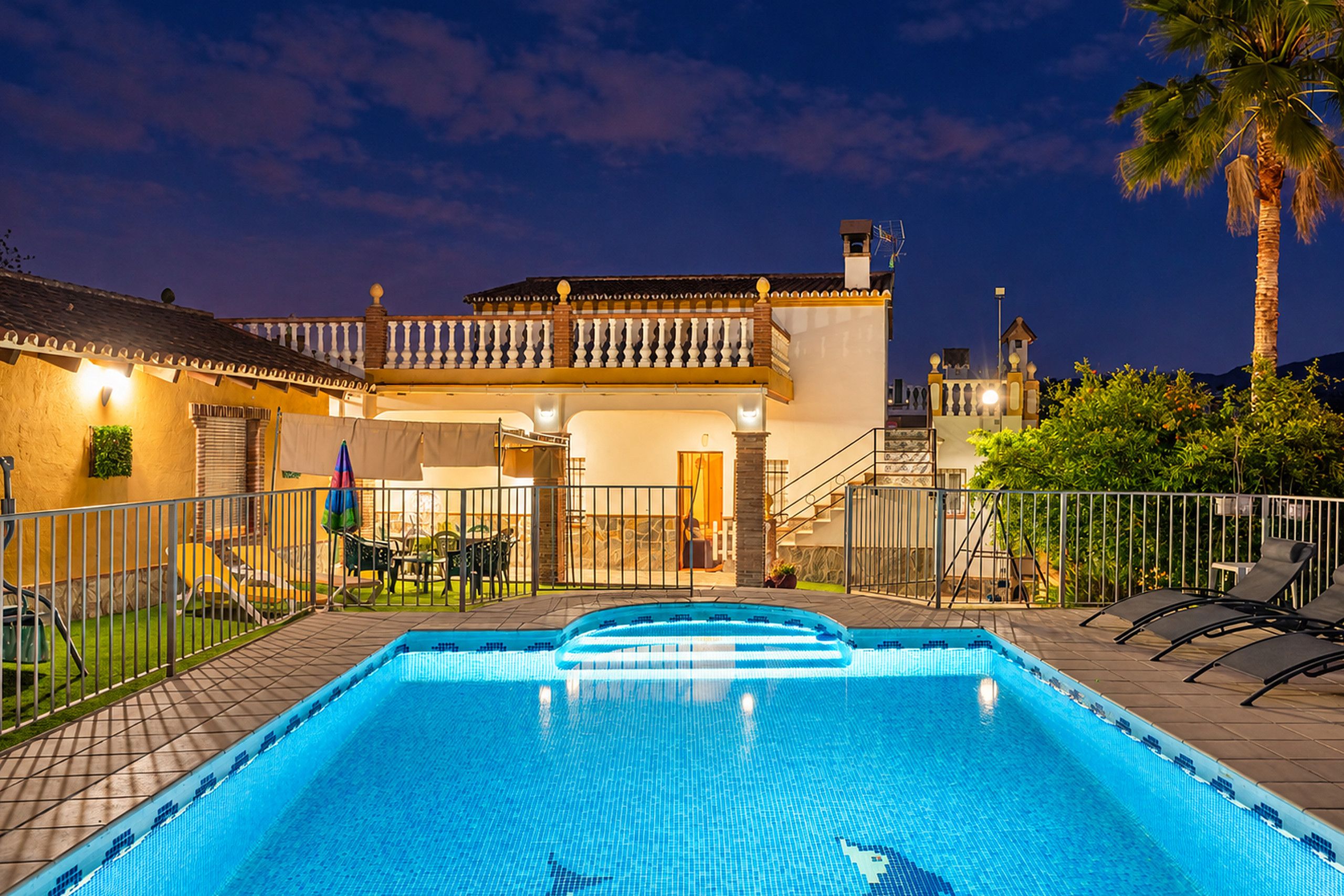 Una piscina con solárium azulejado, un patio cubierto y un edificio de dos plantas con balcón.