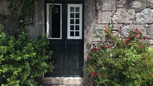 Ein Steinhaus mit einer dunkelgrünen Tür, einem kleinen Fenster und einem Garten mit roten Blüten.