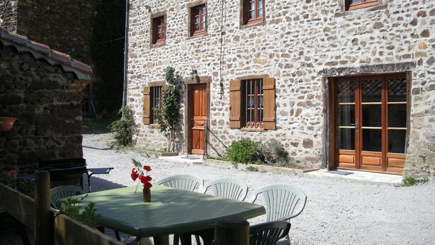 Un edificio de piedra con varias ventanas y puertas, mobiliario exterior con sillas y una mesa, y un pequeño jardín.
