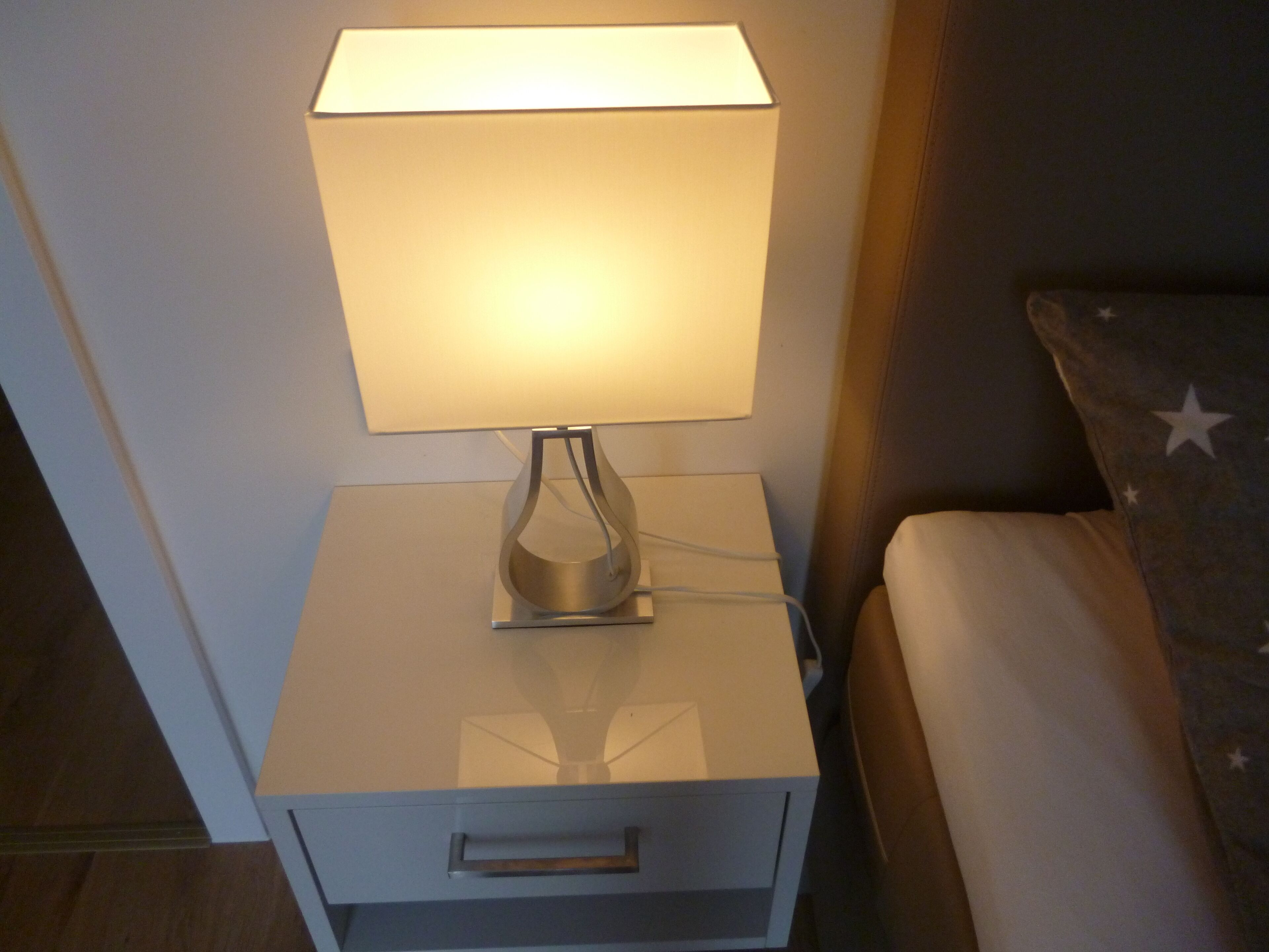 A bedside table with a lamp, a drawer, and a bed with a pillow featuring star patterns.