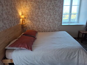 A room with a bed, a wall-mounted lamp, a window, and a chair. - Small gite - beautiful, peaceful setting in an old farmhouse in the heart of the marshes (Terre-Et-Marais)