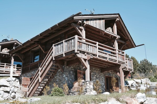 Eine rustikale Holzhütte mit Steinunterbau, einem Holzbalkon und einer Treppe, die zum Obergeschoss führt.