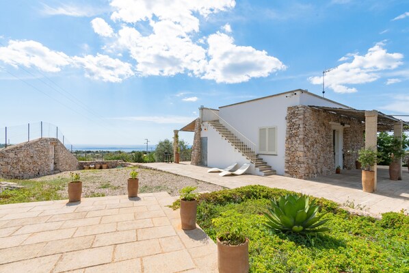 A stone house with a staircase leading to a covered porch, surrounded by potted plants and a clear view of the sea.