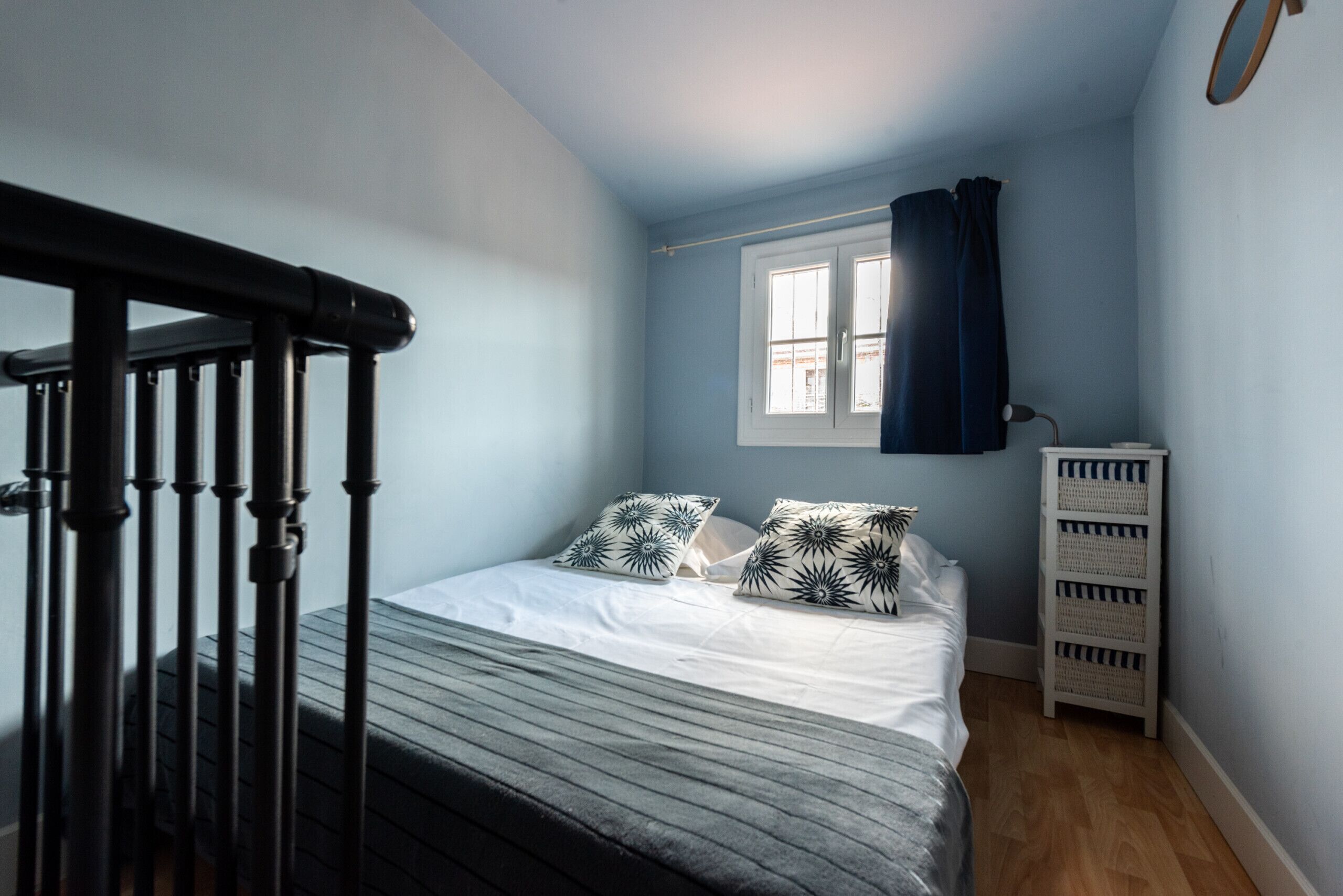 A bedroom with a bed, a window with curtains, a white dresser, and a black railing.