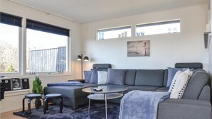 Una sala moderna con un sofá gris, una mesa de centro y dos mesas auxiliares. Hay ventanas grandes con persianas, un cuadro en la pared y un pequeño balcón con plantas.