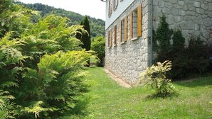A stone building with a stone wall and wooden shutters, surrounded by lush greenery and a hillside in the background.