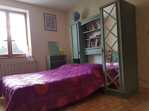 Un dormitorio con colcha morada, una ventana con vistas, un radiador y un armario verde con estantes.
