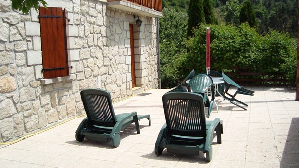 A stone building with a wooden shutter, outdoor seating with green chairs, and a view of a forested hillside.