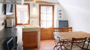 A compact kitchen with a sink, countertop, and wooden cabinets. There is a window with curtains, a small table with chairs, and a television on the counter.