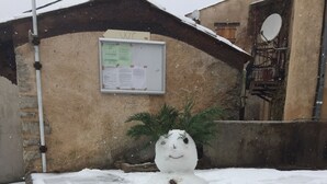 A snowman in a snowy outdoor area with a building in the background.