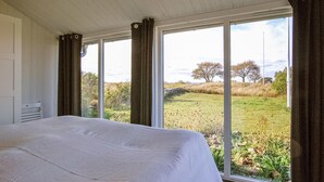 Ein Schlafzimmer mit einem großen Bett, einem Fenster mit Vorhängen und Blick auf ein grasbewachsenes Feld und Bäume.