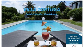 Vue sur la piscine de la Villa Tortuga à Saint-François, en Guadeloupe, avec une table à manger, des boissons et un parasol tropical.