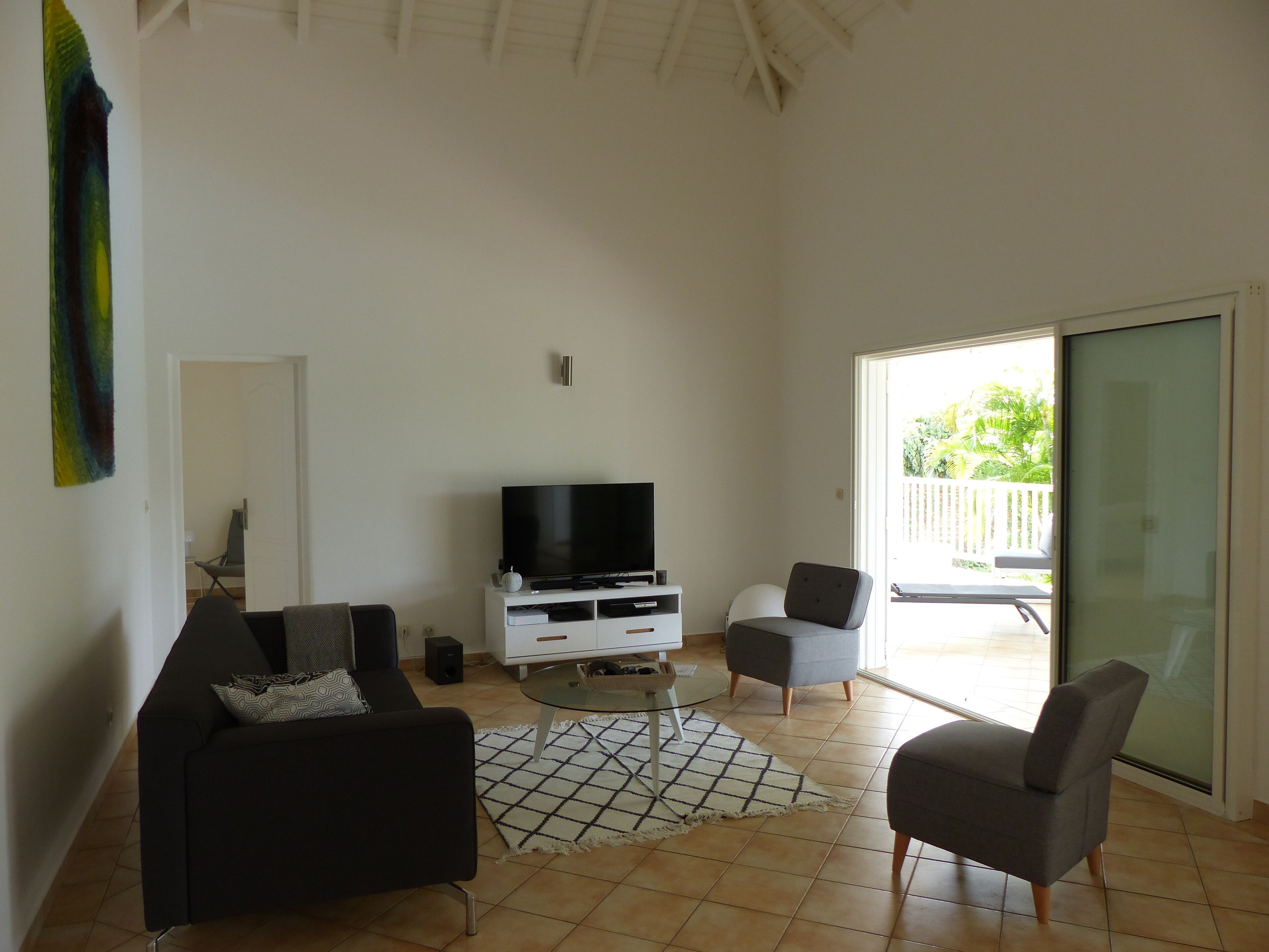 A living room with a flat-screen TV on a white cabinet, two grey armchairs, a black sofa, and a patterned rug.