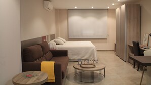 A modern hotel room with a brown sofa, a round coffee table, a bed with white bedding, and a large window.