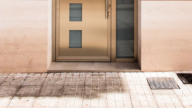 A modern building entrance with a large, beige-colored door featuring a glass panel and a metal handle.