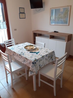 A dining area with a table covered in a floral tablecloth, white chairs, a wooden cabinet, and a mounted television. - "LA MIMOSA" DREAM VILLA near the beach with a beautiful private garden (Capoliveri)