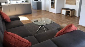 Ein modernes Wohnzimmer mit einem grauen Sofa, roten Kissen, einem Glastisch und einer Küchenzeile mit grauen Schränken und einer Mikrowelle.