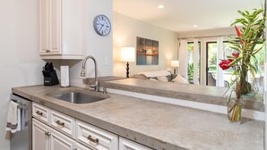 A kitchen with a double sink, white cabinetry, a clock, and a painting on the wall. - Kihei Garden Estates B-103 - Modern Design, Bright & Clean, Concrete Floors, AC! (Kihei)