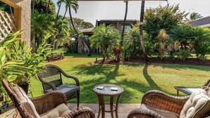 A patio area with wicker furniture, a small table, and a view of a well-maintained lawn and buildings in the background. - Kihei Garden Estates B-103 - Modern Design, Bright & Clean, Concrete Floors, AC! (Kihei)