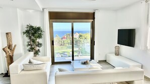 A modern living room with a sofa, a coffee table, and a TV. A large window offers a view of the sea and a garden.