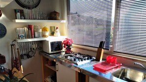 A compact kitchen with a microwave, stove, and sink. There are various kitchen utensils hanging on the wall, and a window with blinds.