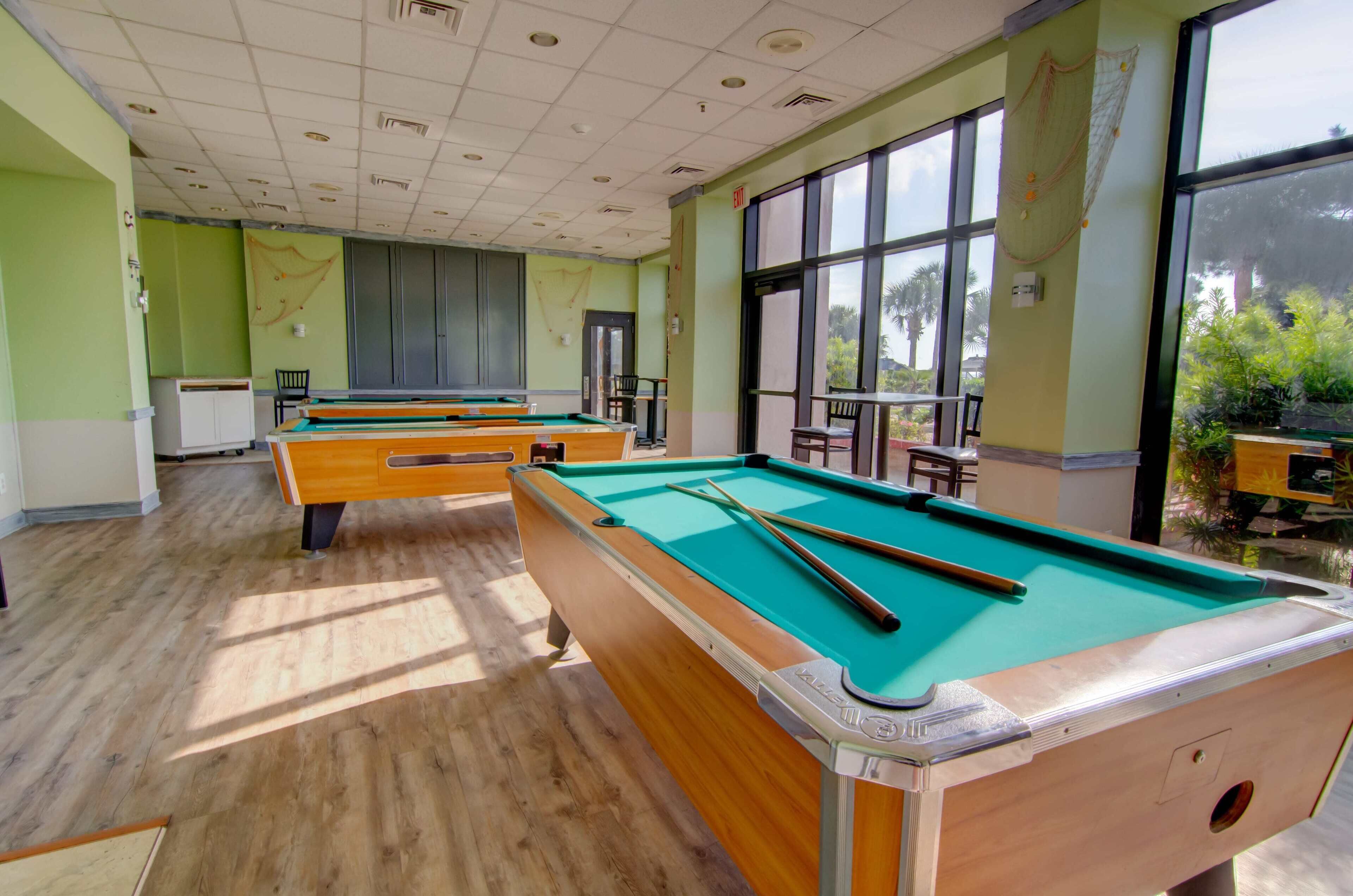 A pool table area with multiple tables, chairs, and a view of greenery outside.