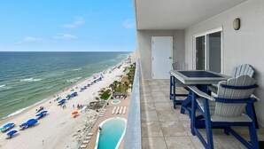 A balcony with a view of a beach and ocean, a pool, and a table with chairs.