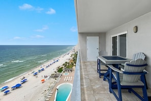 A balcony with a view of a beach and ocean, a pool, and a table with chairs.