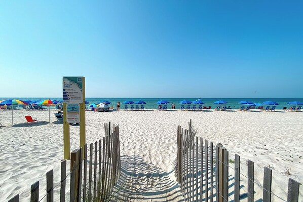 A beach with a wooden fence leading to the water, blue umbrellas, and beach chairs.