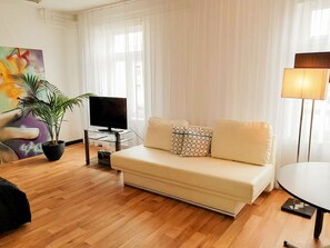 Een moderne woonkamer met een bank, een televisie op een glazen standaard, een potplant en een schilderij aan de muur.