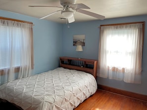 A bedroom with a bed, a ceiling fan, a nightstand, and a window with sheer curtains.