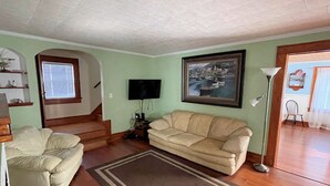 A living room with a beige sofa, a television, a framed cityscape painting, and a staircase leading to another room. - Blue Water - Single Family Home - In Town - LG. FENCEED IN YARD, NO LINENS (Chincoteague)