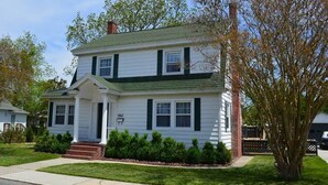 A two-story white house with green shutters, a front porch, and a well-maintained lawn. - Blue Water - Single Family Home - In Town - LG. FENCEED IN YARD, NO LINENS (Chincoteague)