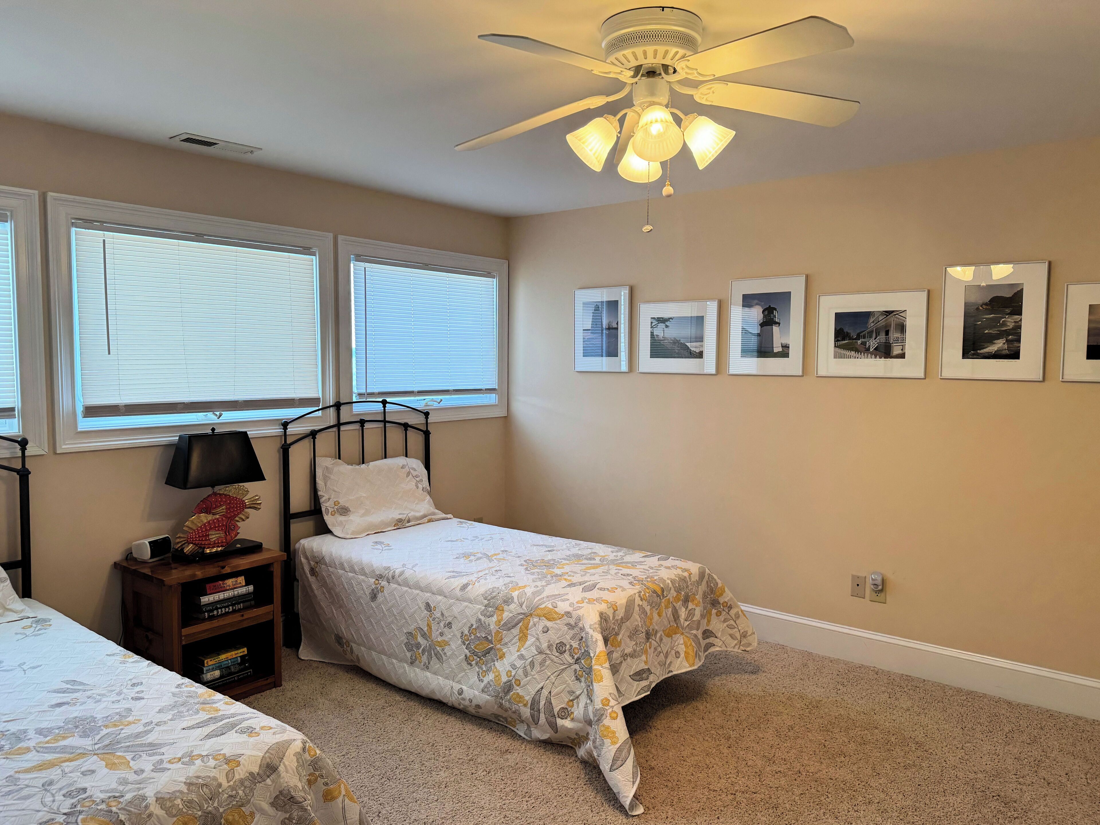 A bedroom with two beds, a ceiling fan, a nightstand with books, and framed pictures on the wall.