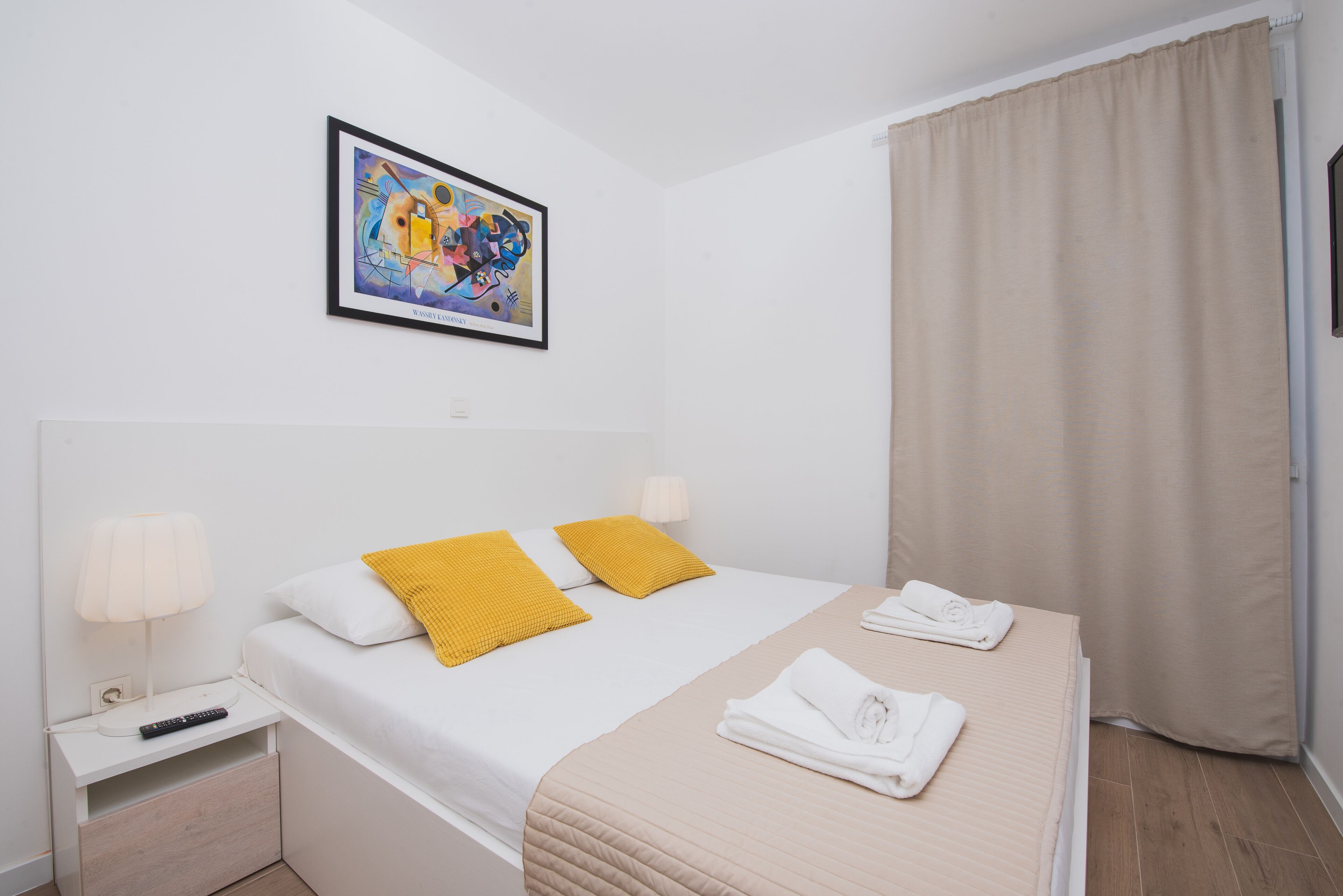 A hotel room with a bed, two pillows, a nightstand with a lamp, a framed picture on the wall, and a window with curtains.