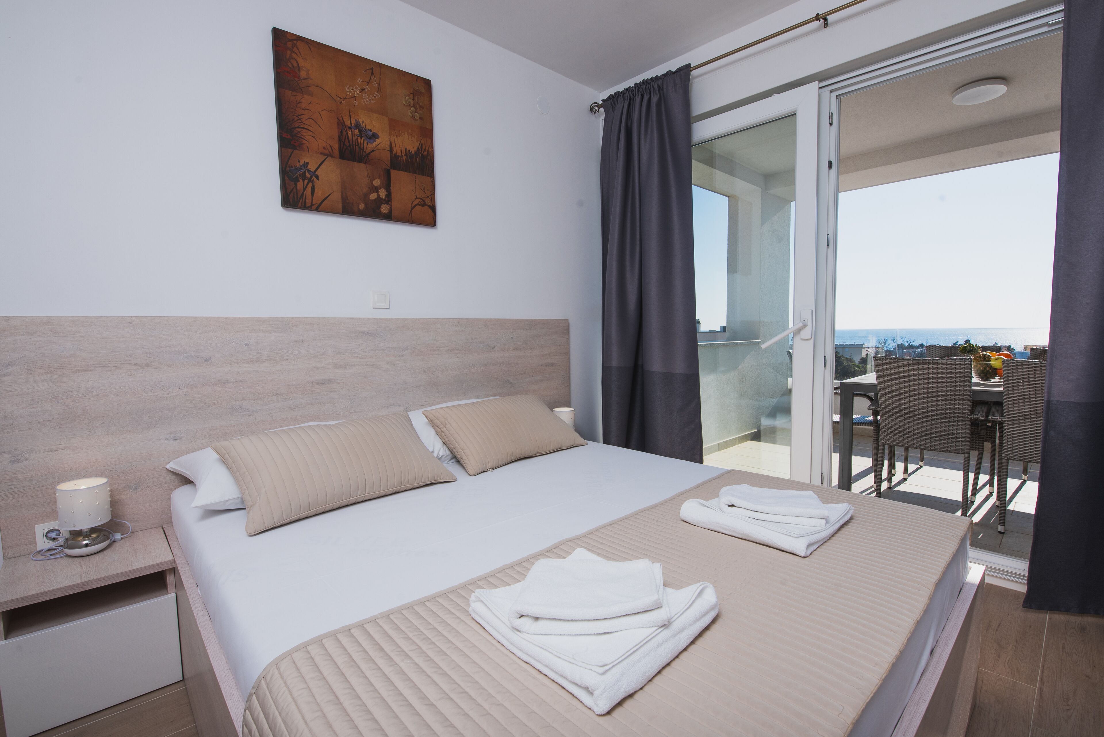 A hotel room with a bed, a nightstand, a painting, and a balcony with a table and chairs.