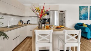A modern kitchen with white cabinets, a wooden countertop, and a central island with chairs. There is a vase with flowers on the island, and a blue couch in the background. - Villa Festina Lente in Makarska, heated pool (Makarska)