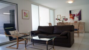 Ein Wohnzimmer mit braunem Ledersofa, einem Glastisch, einem Korbstuhl und einem Essbereich mit einem weißen Schrank und einem Blumenvasen.