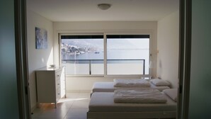 Ein Schlafzimmer mit zwei Betten, einem Schminktisch und einem großen Fenster mit Blick auf einen See und Berge.