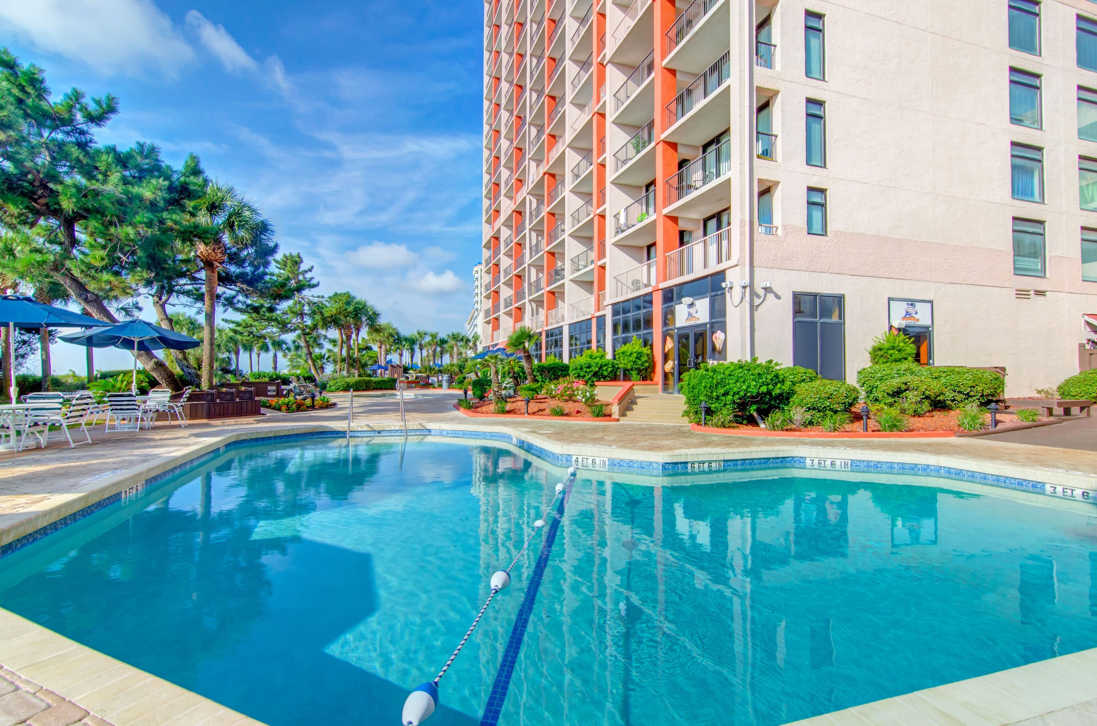A swimming pool with lounge chairs and umbrellas, surrounded by a building with balconies and a clear sky.