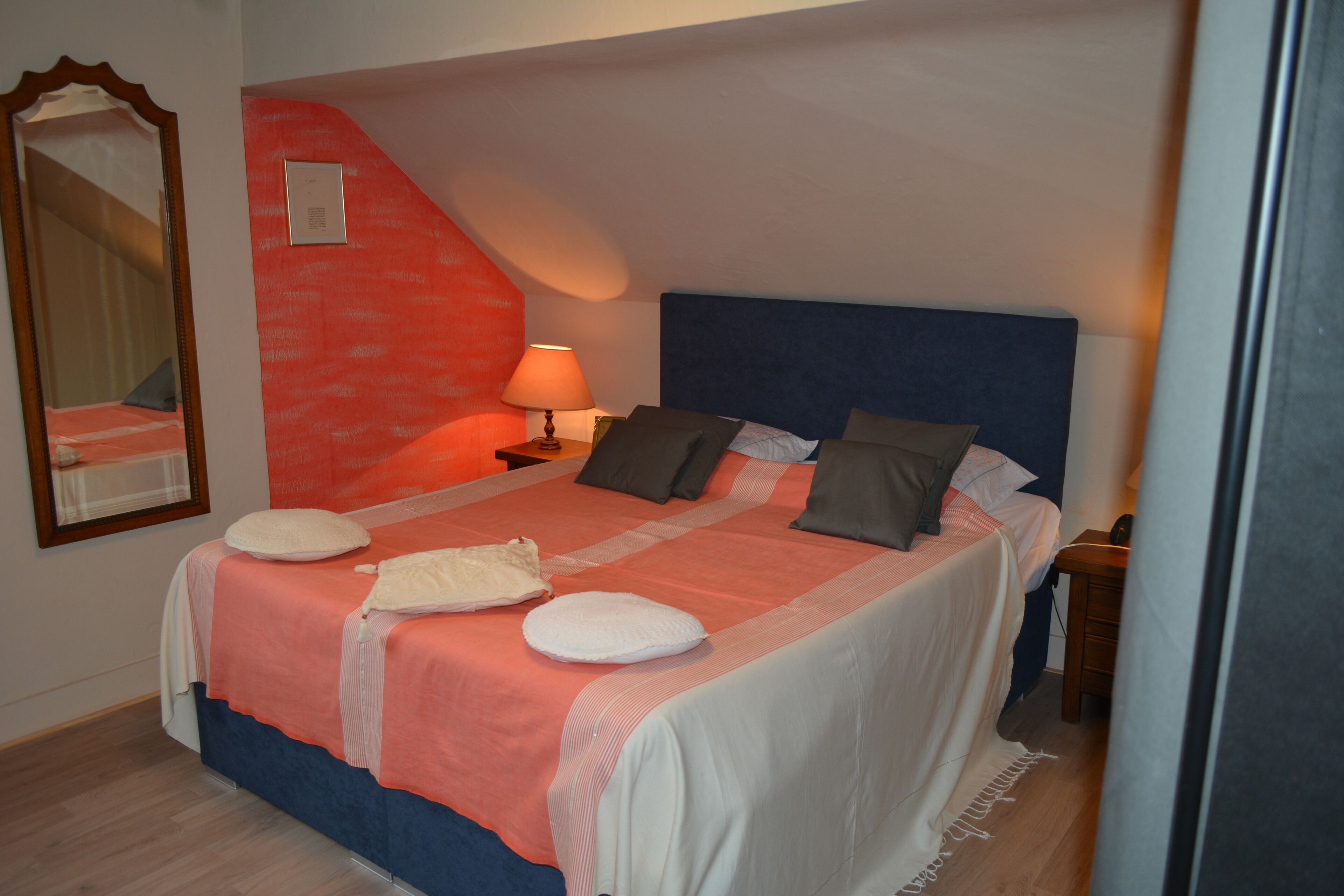 Ein Schlafzimmer mit einem Bett, Kissen, einem Nachttisch, einer Lampe, einem Spiegel und einer roten Akzentwand.