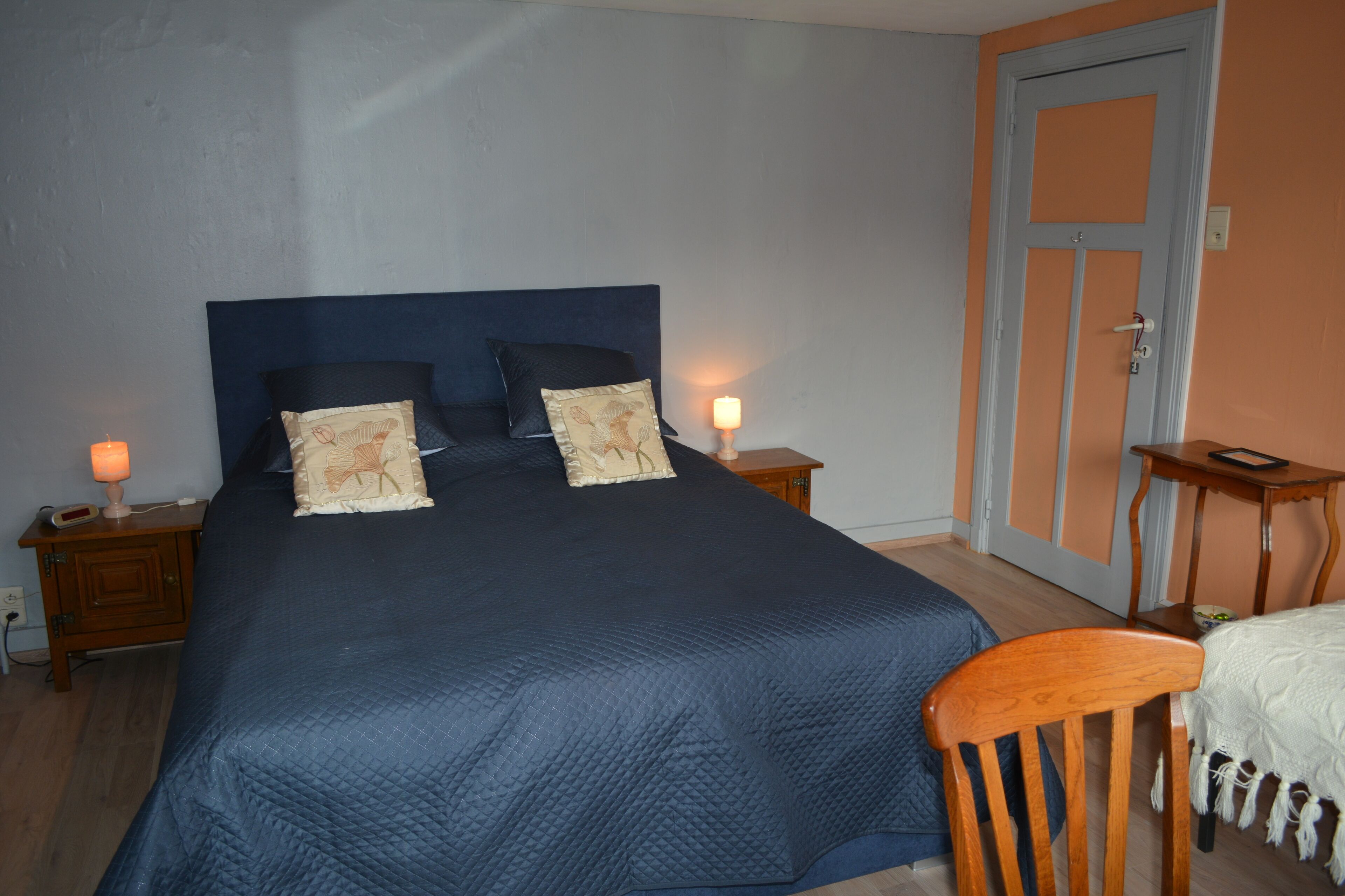 Un dormitorio con una cama grande, dos mesitas de noche, una mesita auxiliar, una silla de madera y una puerta.