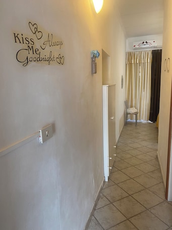 A hallway with a wall sign saying "Kiss Me Goodnight," a light fixture, a door with a curtain, and a tiled floor.