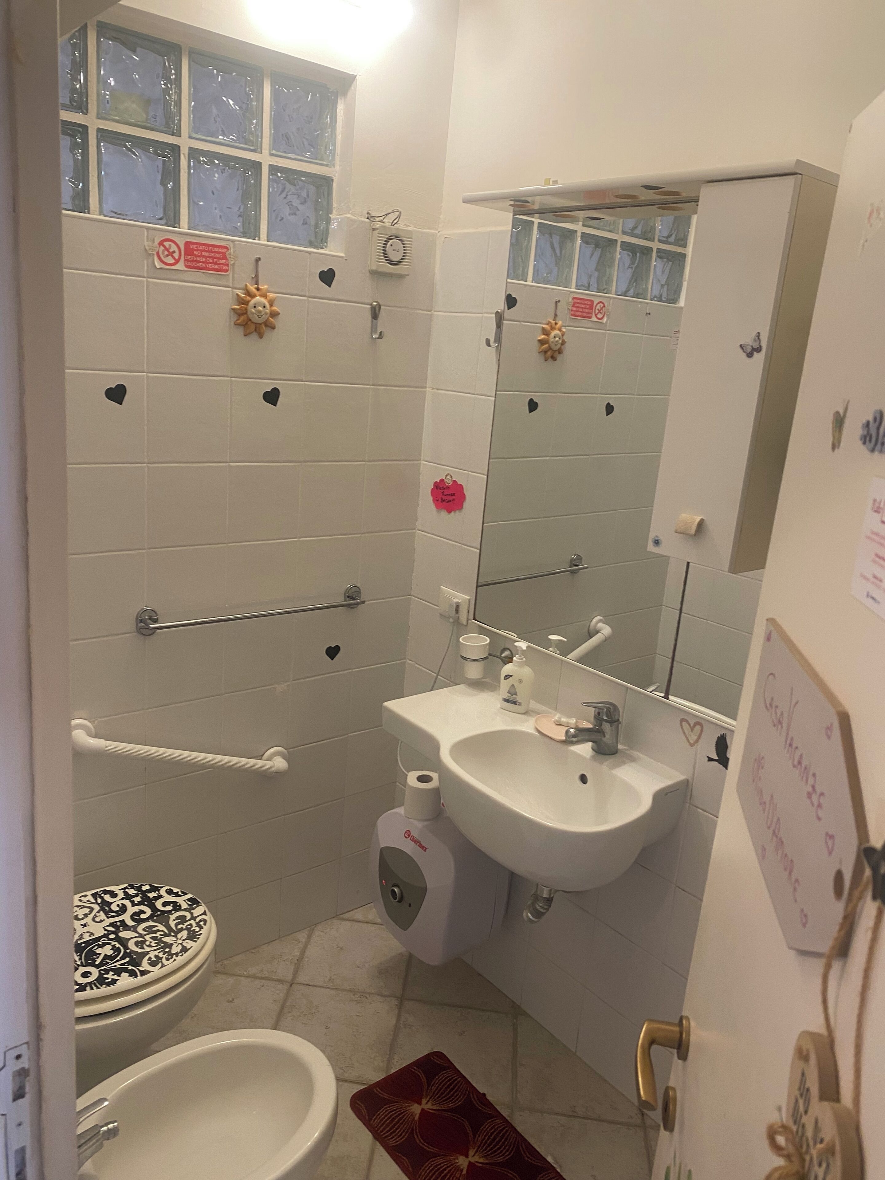 Ein kleines Badezimmer mit Toilette, Bidet, Waschbecken und Spiegel. An den Wänden befinden sich Handläufe und über der Toilette ein Fenster.