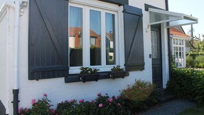 Un bâtiment blanc avec des volets sombres, une porte et une fenêtre donnant sur une maison.