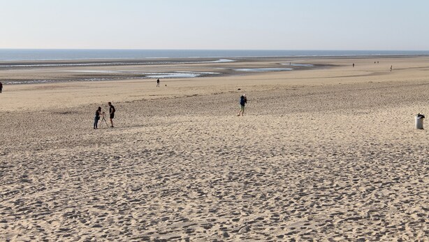 Une vaste plage de sable où l’on voit des gens se promener et rester debout.
