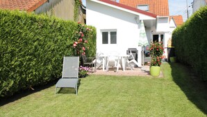 Une maison blanche avec un toit rouge, entourée d’un jardin bien entretenu et d’une haie.