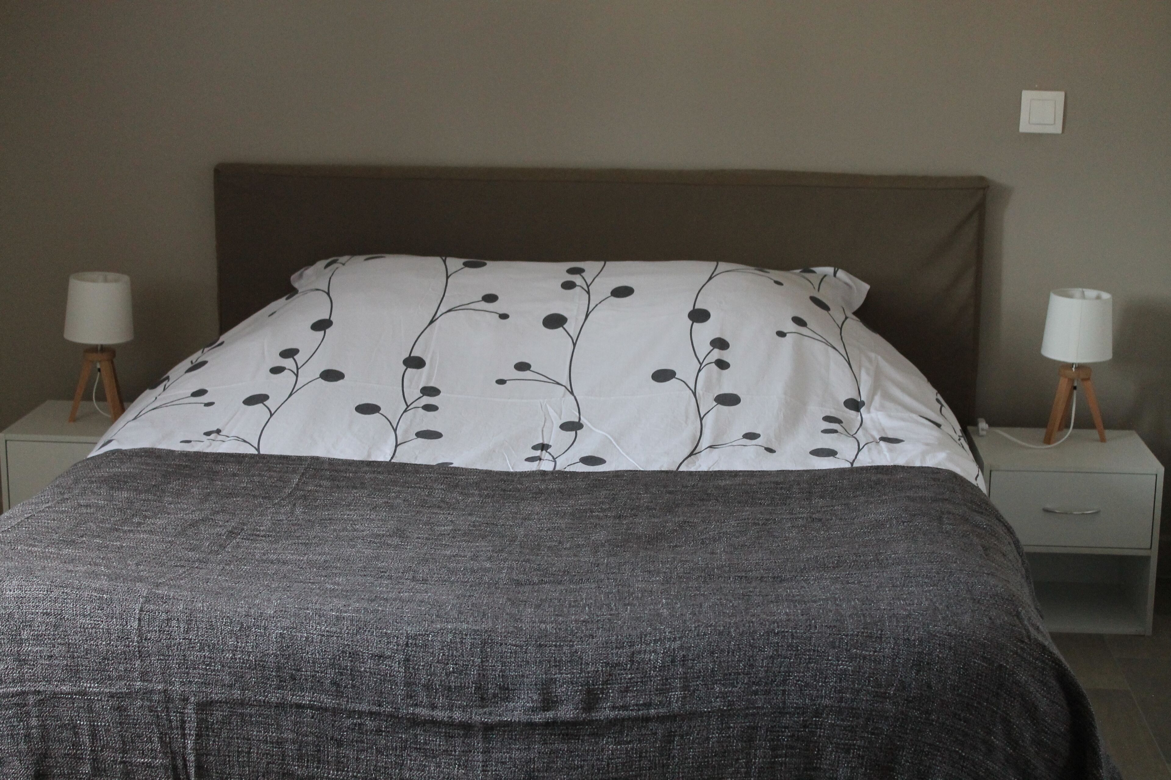 Un lit bien fait, recouvert d’une couvre-lit à motifs floraux noirs et blancs, deux tables de chevet avec des lampes, et un mur sobre.