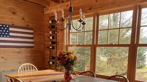 Une salle à manger en bois avec une grande table, des chaises, un lustre, un drapeau et une fenêtre donnant sur des arbres.