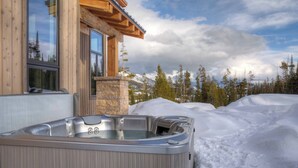 Ein Whirlpool vor einer Holzhütte mit Steinunterbau, umgeben von Schnee und Bäumen.